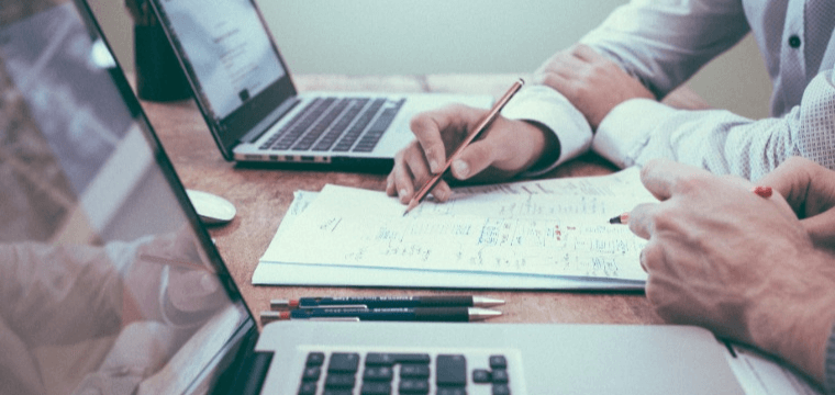 Image of people writing on a notepad, with two laptops beside them. Depicting Lifebit's approach to secure collaboration and safeguarding data integrity